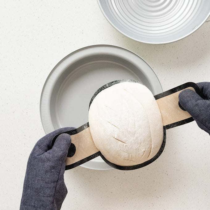 Pair of gloved hands holding shaped sourdough dough on a wooden peel above a gray mixing bowl with a proofing basket nearby, part of a handcrafted beginner baking kit in Baking Supplies, Kitchen & Food collections.