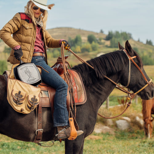 Woman wearing a cowboy hat and casual jacket sitting on a dark brown horse with a saddle bag featuring a cast iron grill pan, set against a grassy outdoor landscape with hills, perfect for Kitchen & Food and USA Made collections.