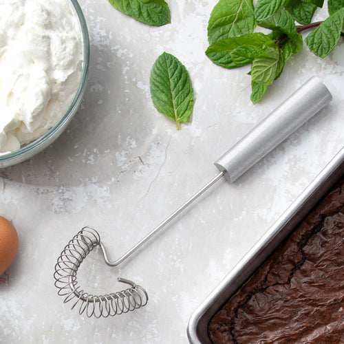 Rada Handi-Stir whisk with a stainless steel coil designed to reach corners and scrape pans, shown with fresh mint leaves, an egg, whipped cream, and brownies, fitting Kitchen & Food and USA Made collections.