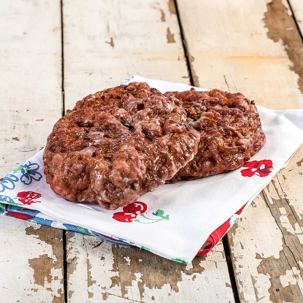 Two freshly baked fruit fritters with a sweet glaze resting on a colorful floral cloth, showcasing a handmade treat perfect for Kitchen & Food, Snacks and Sweets, USA Made, Holiday Hosting, and Best Sellers collections.