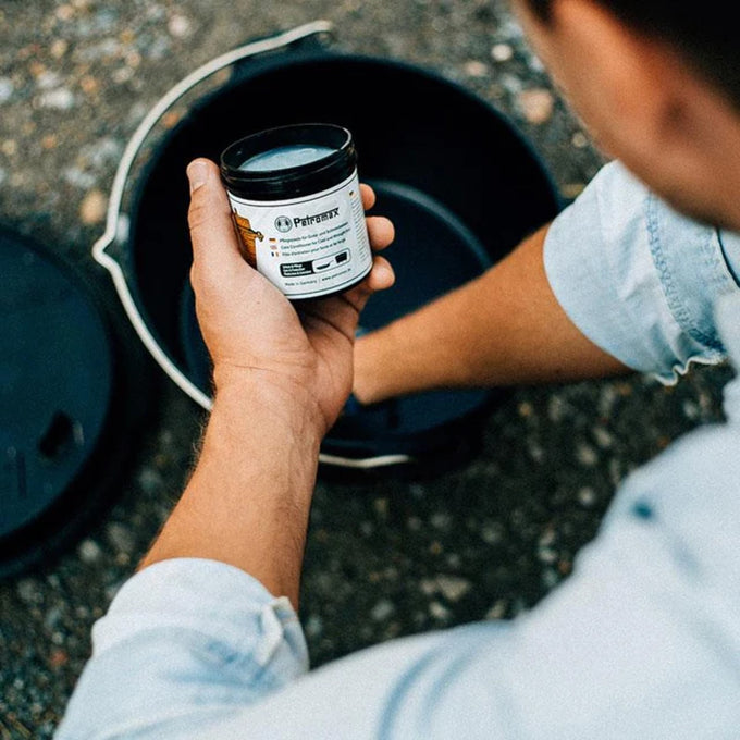 Person holding a small container of Petromax Care Conditioner for Cast and Wrought Iron above a Dutch oven outdoors, ideal for Kitchen & Food, Cookware, Cast Iron, Best Sellers, and Promo Eligible collections.