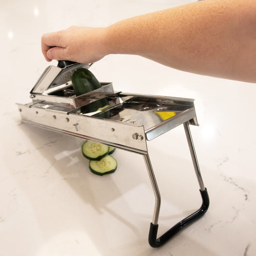Handheld stainless steel mandoline veggie slicer with foldable legs and safety holder, slicing cucumber on a white countertop, part of Kitchen & Food and Food Prep collections.