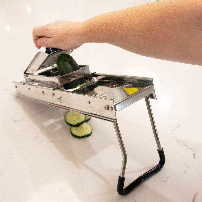 Handheld stainless steel mandoline veggie slicer with foldable legs and safety holder, slicing cucumber on a white countertop, part of Kitchen & Food and Food Prep collections.