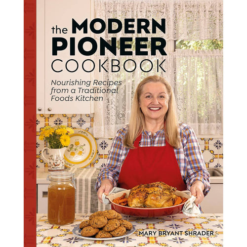 Smiling woman in a checkered shirt and red apron holding a roasted chicken on a red platter, standing in a bright traditional kitchen with cookies, broth jar, and vintage decor, part of handcrafted Amish made cookbooks in the Books and Best Sellers collections.