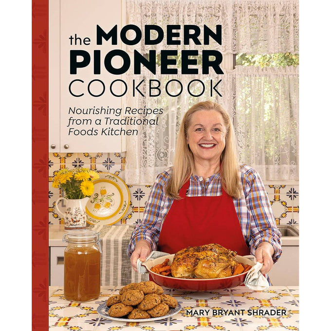 Smiling woman in a checkered shirt and red apron holding a roasted chicken on a red platter, standing in a bright traditional kitchen with cookies, broth jar, and vintage decor, part of handcrafted Amish made cookbooks in the Books and Best Sellers collections.