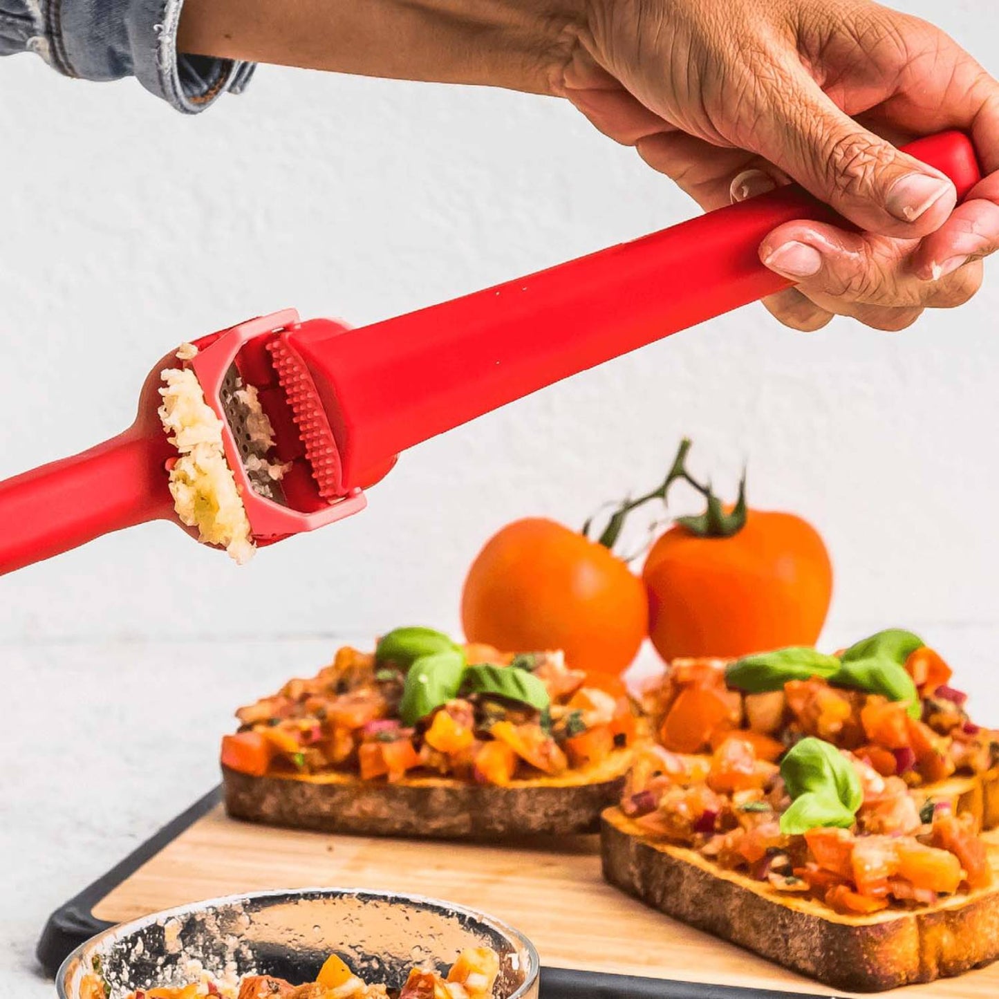 Red garlic press in use mincing unpeeled cloves with peel ejecting feature, shown over bruschetta with tomatoes and basil, suitable for Kitchen & Food, Food Prep, and Kitchen Gadgets collections.