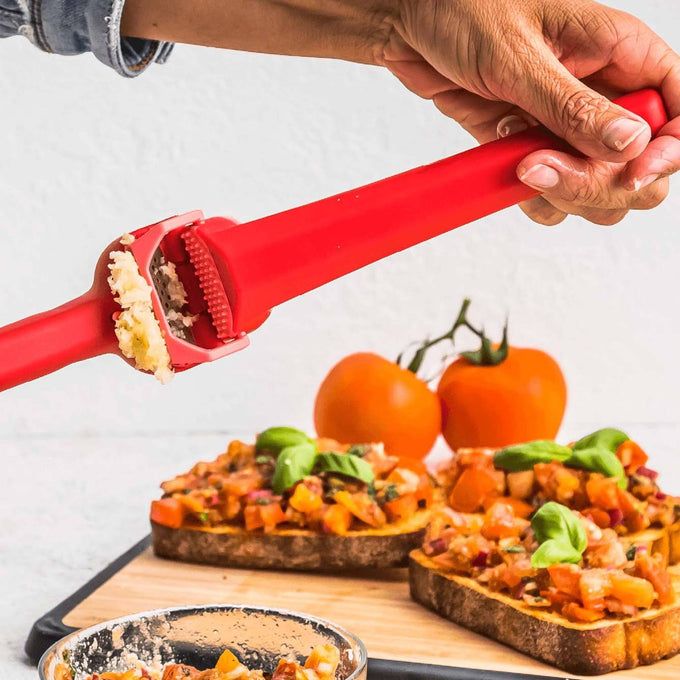 Red garlic press in use mincing unpeeled cloves with peel ejecting feature, shown over bruschetta with tomatoes and basil, suitable for Kitchen & Food, Food Prep, and Kitchen Gadgets collections.
