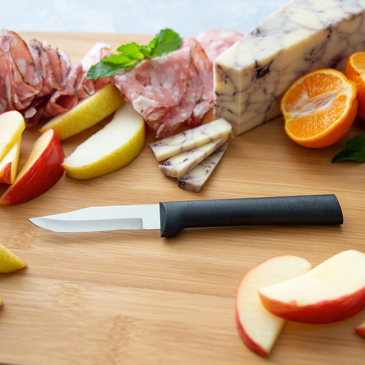 Paring knife with a non-serrated stainless steel blade and smooth black handle on a wooden board with sliced apples, pear, cured meats, marbled cheese, and mandarin oranges, USA made.