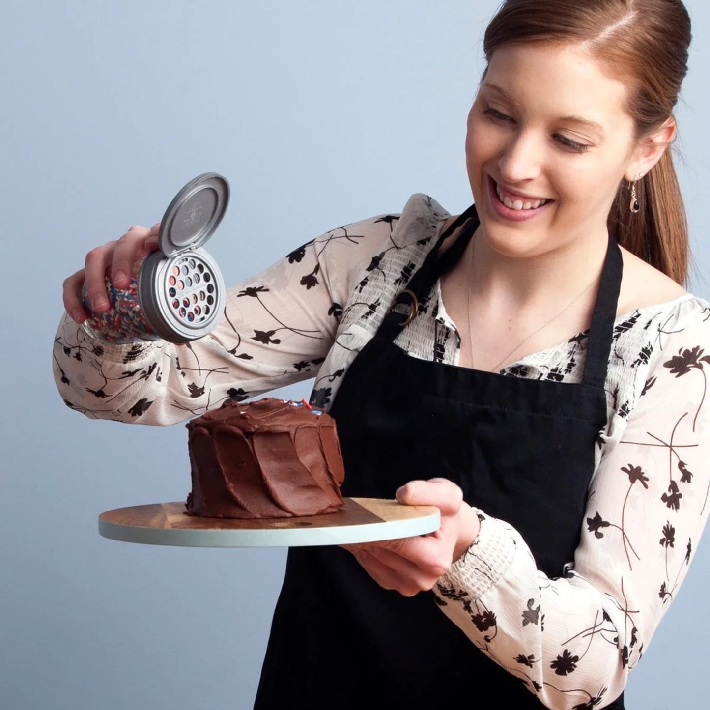 Smiling woman wearing black apron sprinkling spices from a regular-mouth Mason jar with a flip shaker lid onto a chocolate frosted cake, highlighting easy kitchen gadget use for cooking helpers.