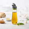 Clear glass oil bottle with automatic flip-top spout and drip catcher, designed for mess-free pouring, featured with fresh bread, basil, and garlic in a bright kitchen setting.
