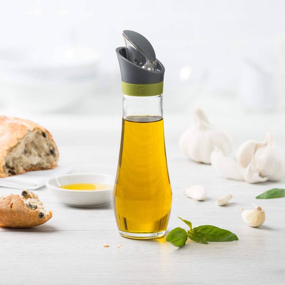 Clear glass oil bottle with automatic flip-top spout and drip catcher, designed for mess-free pouring, featured with fresh bread, basil, and garlic in a bright kitchen setting.