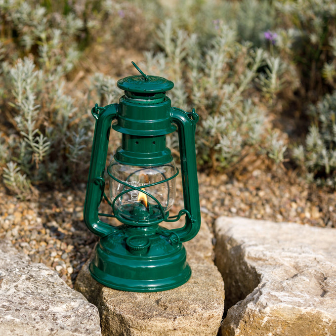 Handcrafted green galvanized oil lantern with thermal heat-resistant SUPRAX globe, glowing gently while resting on a stone surface outdoors, ideal for Lamps & Lighting, Camping, and Emergency Supplies collections.