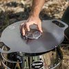 Hand placing a round metal insert with concentric cut-out design into the center of a dark Atago Griddle Plate on a shiny stainless steel stove, ideal for Farm, Garden & Tool, Stoves, Camping, Grilling, and Best Sellers collections.