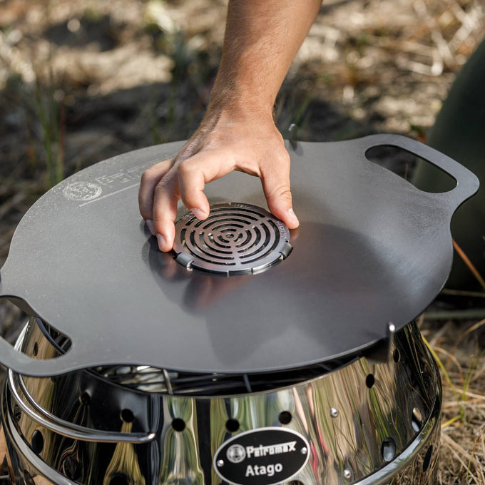 Hand placing a round metal insert with concentric cut-out design into the center of a dark Atago Griddle Plate on a shiny stainless steel stove, ideal for Farm, Garden & Tool, Stoves, Camping, Grilling, and Best Sellers collections.