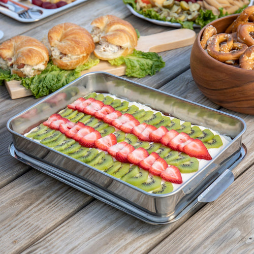 Rectangular stainless steel cake pan filled with a creamy dessert topped with sliced kiwi and strawberries, surrounded by sandwich sliders and a bowl of pretzels on a rustic wooden table.