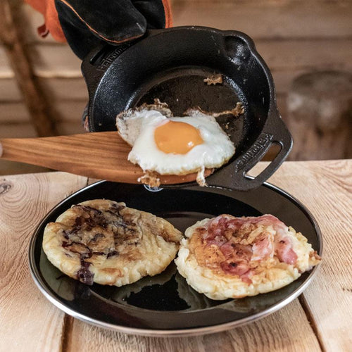 Small handcrafted cast iron skillet with two side handles being used to cook a fried egg, paired with a plate holding two hearty flatbreads, perfect for camping and kitchen use collections.
