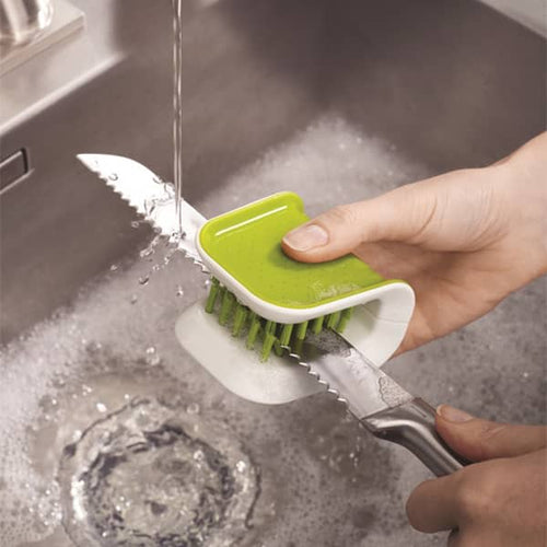 Handheld green and white dual-sided brush cleaning a serrated knife under running water in a stainless steel sink, featured in kitchen accessories and cleaning utensils collections.