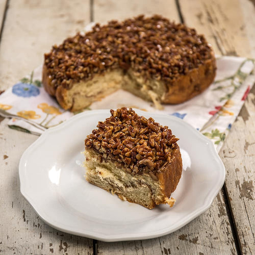 Slice of homemade pecan roll topped with chopped nuts and caramel glaze, handcrafted in the USA, displayed on a white plate with the full roll in the background on rustic wood.
