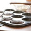 Carbon steel whoopie pie pan with six freshly baked chocolate cakes rising evenly in shallow cavities, a glass bowl of white frosting and wooden spatula resting nearby, ideal for Kitchen & Food, Cookware, Baking Supplies, Best Sellers, and Promo Eligible collections.