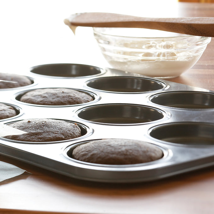 Carbon steel whoopie pie pan with six freshly baked chocolate cakes rising evenly in shallow cavities, a glass bowl of white frosting and wooden spatula resting nearby, ideal for Kitchen & Food, Cookware, Baking Supplies, Best Sellers, and Promo Eligible collections.
