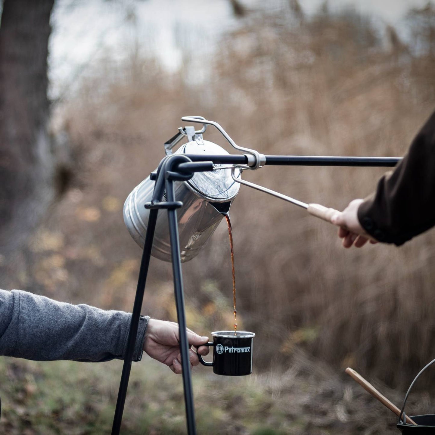 Pouring hot coffee from a campfire percolator using a metal pouring aid with wooden handle, held over a black enamel mug outdoors, ideal for Farm, Garden & Tool, Camping, and Best Sellers collections.