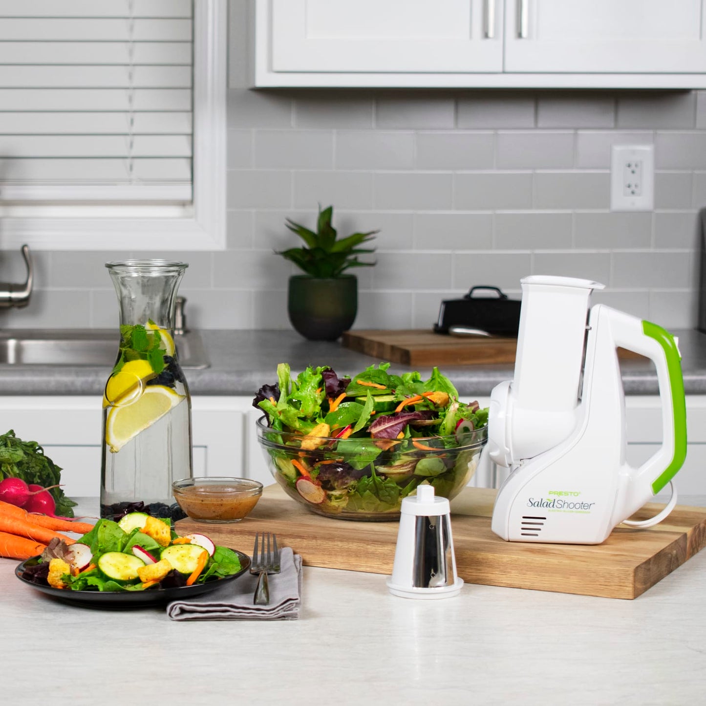 Electric salad slicer with white and green plastic body displayed on kitchen countertop with fresh mixed salad, carrots, dressing bowl, and glass water pitcher, featured in Kitchen & Food, Food Prep, and Kitchen Gadgets collections.