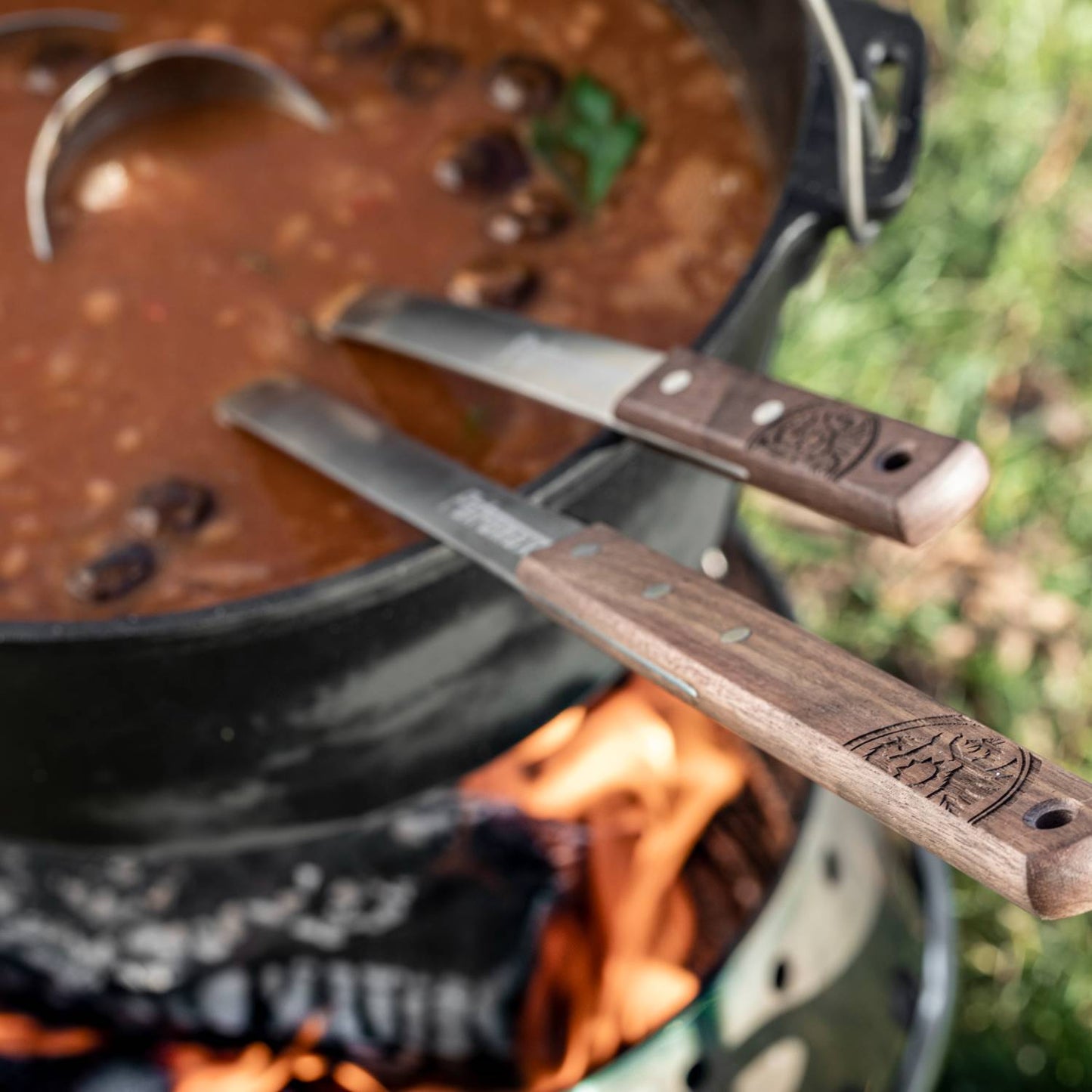 Cast iron pot filled with hearty bean soup cooking over an open campfire, accompanied by wooden-handled utensils resting on the pot, perfect for outdoor cooking in camping and farm collections.