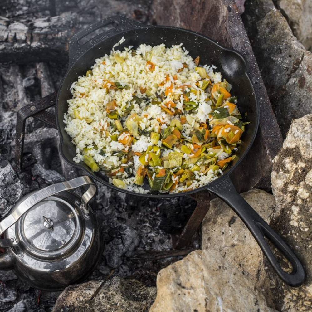 Cast iron skillet with a long handle cooking a vegetable and rice dish over an open campfire, surrounded by rocks and accompanied by a metal kettle, ideal for Farm, Garden & Tool, Camping, and Cookware collections.