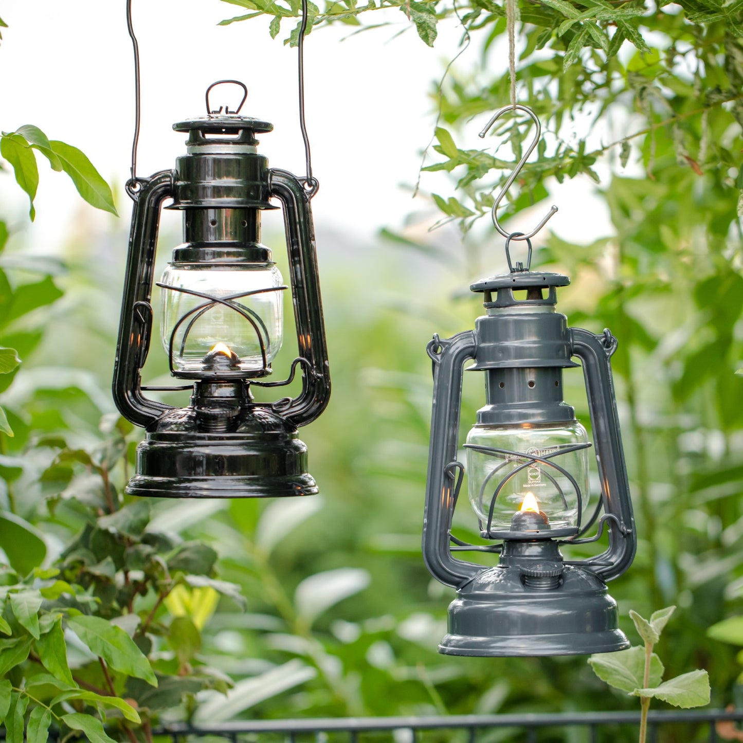 Two handcrafted galvanized oil lanterns, one black and one dark gray, softly glowing while hanging outdoors among lush green foliage, ideal for camping and emergency supplies collections.