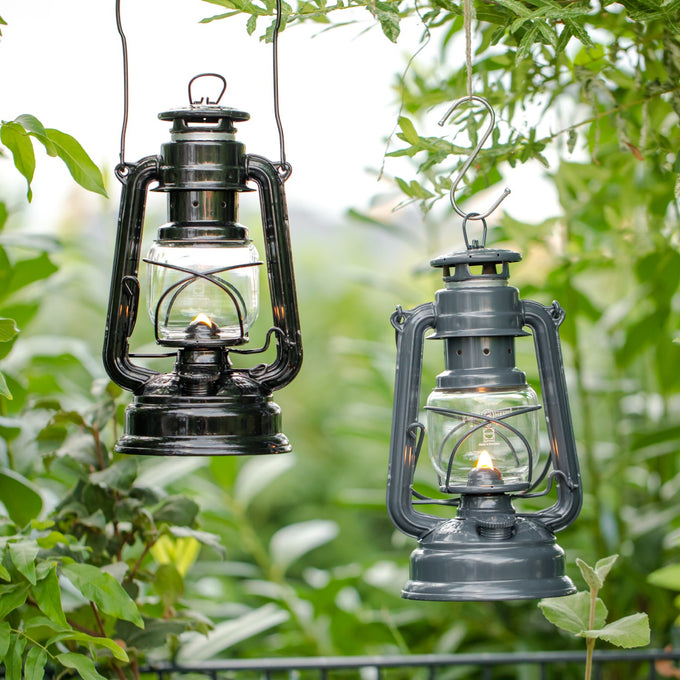 Two handcrafted galvanized oil lanterns, one black and one dark gray, softly glowing while hanging outdoors among lush green foliage, ideal for camping and emergency supplies collections.