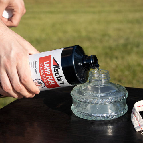 Hand pouring clear Aladdin lamp fuel from a black 32 oz bottle labeled no kerosene odor into a glass lamp reservoir outdoors, ideal for Lamps & Lighting and Lamp and Lantern Fuel collections.