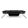 Black steel portable fire pit with angled stainless steel legs and an attached baking compartment, designed for versatile outdoor cooking in camping and garden settings.