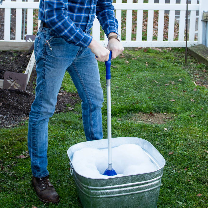 Person using a collapsible hand-operated laundry washer plunger in a galvanized metal tub filled with soapy water outdoors, featured in Home & Decor and Laundry Supplies collections.