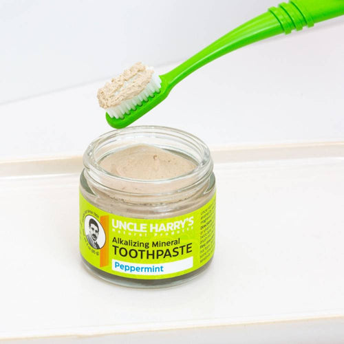 Small glass jar of handcrafted alkalizing peppermint mineral toothpaste with bright yellow label, next to a green toothbrush holding a scoop of fresh paste, promoting Health & Wellness, Grooming and Personal Hygiene, USA Made, and Best Sellers collections.