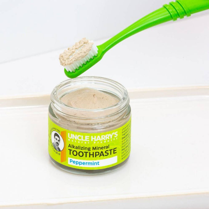 Small glass jar of handcrafted alkalizing peppermint mineral toothpaste with bright yellow label, next to a green toothbrush holding a scoop of fresh paste, promoting Health & Wellness, Grooming and Personal Hygiene, USA Made, and Best Sellers collections.