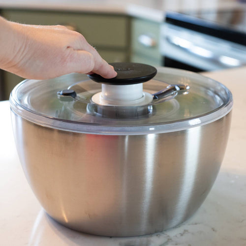Person pressing black push-button handle on brushed stainless steel salad spinner with locking clear lid on kitchen countertop, perfect for Kitchen & Food and Best Sellers collections.