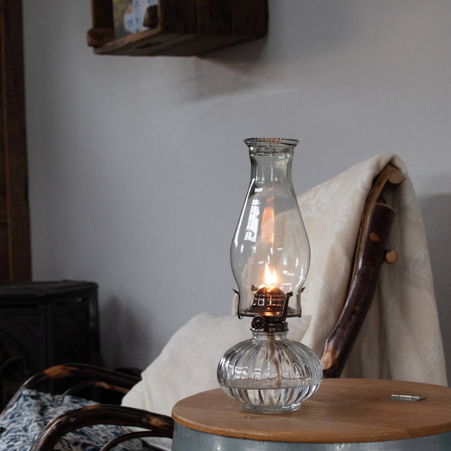 Clear glass oil lamp with a softly glowing flame, featuring an elliptical base and tall chimney, placed on a rustic wooden table in a cozy farmhouse setting, ideal for Farm, Garden & Tool and Emergency Supplies collections.
