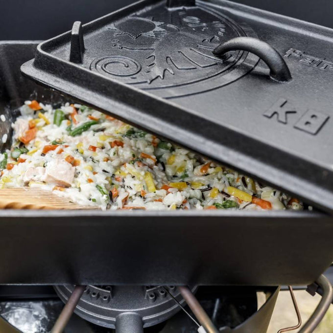 Close-up of a durable cast iron loaf pan with embossed lid cooking a hearty vegetable and rice casserole over a portable camp stove, ideal for camping, kitchen, and farm collections.