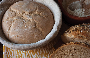 Handcrafted rattan bread proofing basket with a removable cloth liner holding rising dough next to sliced multigrain bread and a bowl of flour, ideal for Kitchen & Food and Baking Supplies.