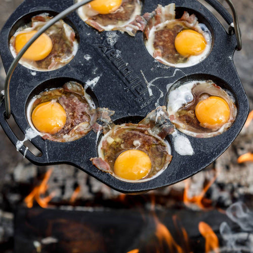 Cast iron muffin pan with six sections cooking eggs and bacon over an open flame, showcasing durable cookware perfect for Farm, Garden & Tool, Camping, Kitchen & Food, Cast Iron, and Best Sellers collections.