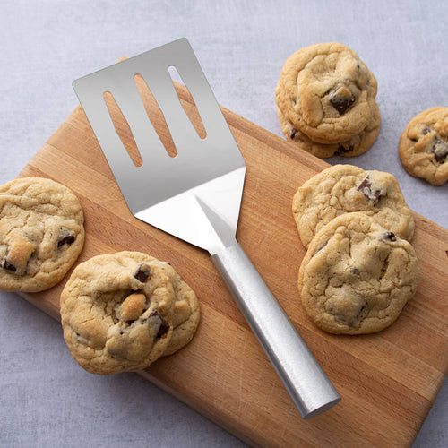 Sturdy metal spatula with slots and angled face for easy flipping and draining, placed on a wooden cutting board with freshly baked chocolate chip cookies, ideal for Kitchen & Food and USA Made collections.