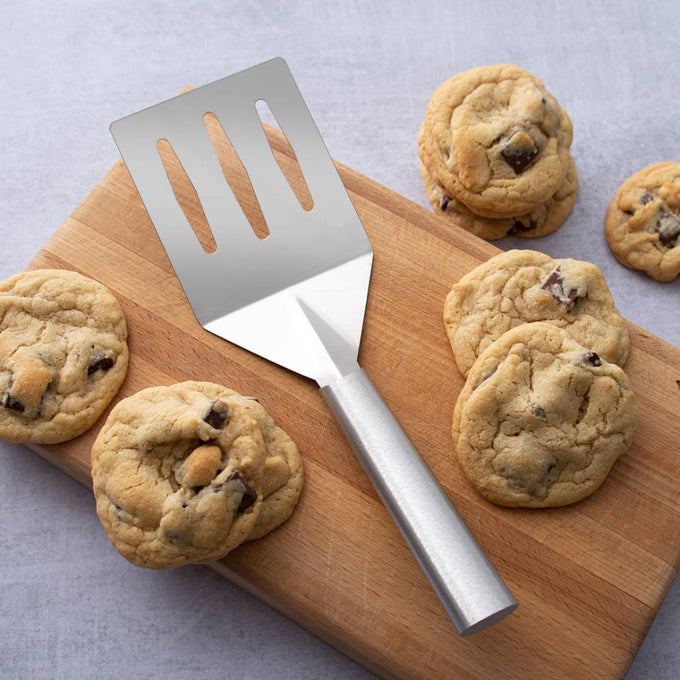 Sturdy metal spatula with slots and angled face for easy flipping and draining, placed on a wooden cutting board with freshly baked chocolate chip cookies, ideal for Kitchen & Food and USA Made collections.