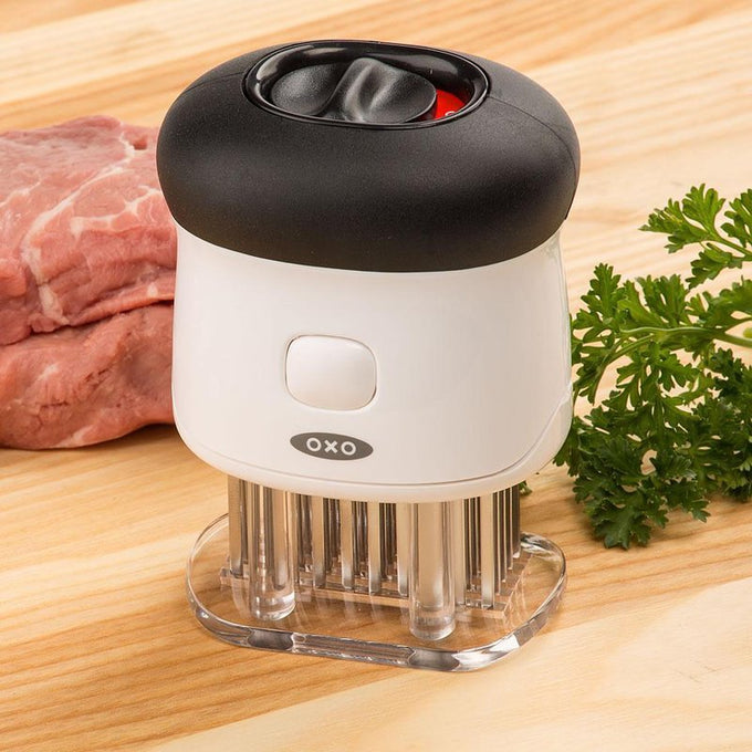 Small white and black meat tenderizer with 58 stainless steel blades locked in clear cover, featuring non-slip grip and safety lock, displayed on wooden surface with raw pork and green parsley, ideal for Kitchen & Food, Specialty Equipment, Butchering Supplies, Best Sellers, and Promo Eligible collections.