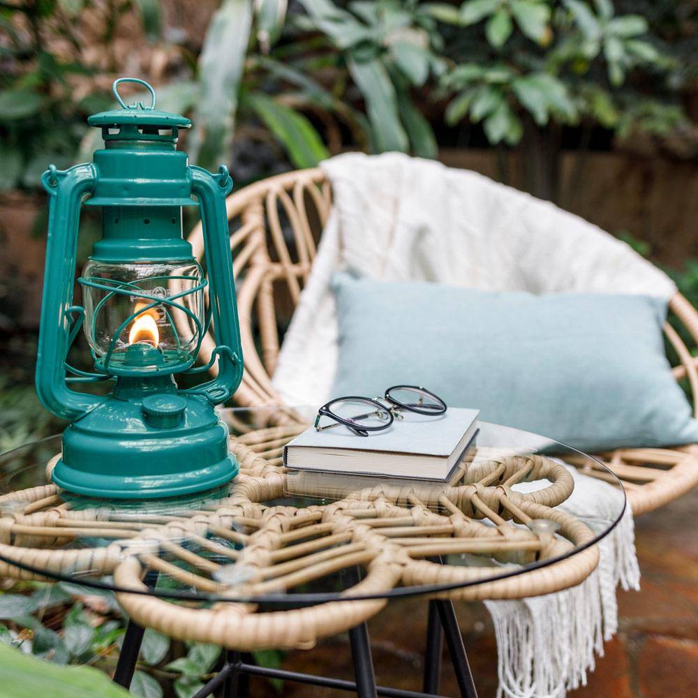 Handcrafted teal galvanized oil lantern with thermal heat-resistant globe softly glowing on a glass-topped wicker table next to books and glasses, set in a cozy outdoor seating area, perfect for Camping, Lamps & Lighting, and Emergency Supplies collections.