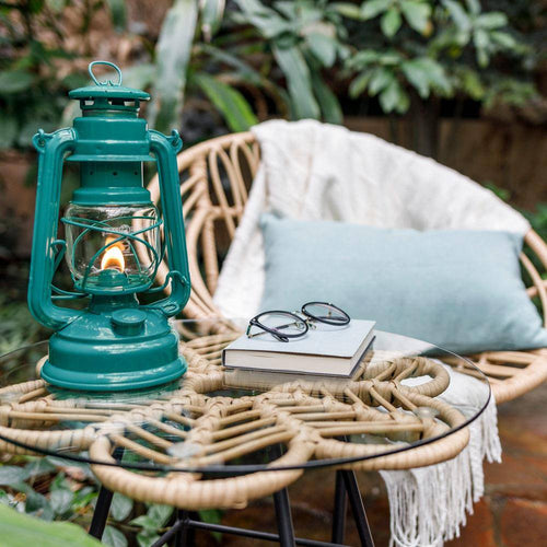 Handcrafted teal galvanized oil lantern with thermal heat-resistant globe softly glowing on a glass-topped wicker table next to books and glasses, set in a cozy outdoor seating area, perfect for Camping, Lamps & Lighting, and Emergency Supplies collections.