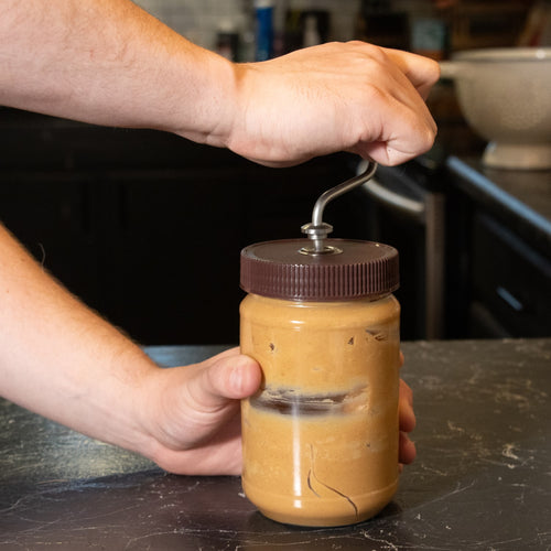 Hand turning a metal stirrer with molded knob attached to a lid on a jar of separated peanut butter, designed to mix oil and prevent messes, made in USA, for Kitchen & Food collections.