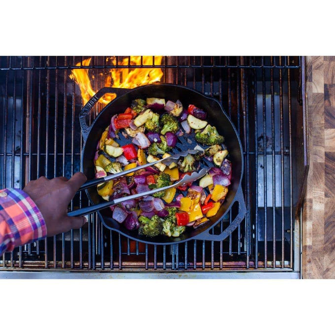 Handcrafted cast iron grill pan with dual handles, filled with colorful vegetables including broccoli, peppers, and onions being stirred over open flame, perfect for Kitchen & Food, Cookware, Cast Iron, Grilling, and Best Sellers collections.