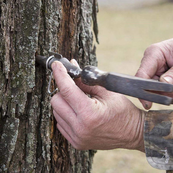 Close-up of hands using a handcrafted solid cast iron spout driver to tap a metal spout into a tree, featured in Farm, Garden & Tool and Maple Syrup Sugaring Supplies collections.