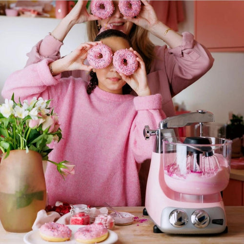 Pink stand mixer with dual control dials mixing pink batter on a kitchen counter surrounded by donuts and flowers, featured in Kitchen & Food, Baking Supplies, and Kitchen Gadgets collections.