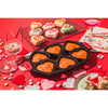 Seasoned cast iron mini cake pan with six heart-shaped cavities filled with baked golden mini cakes on a red table surrounded by conversation heart cookies and festive decorations, featured in Kitchen & Food, Cookware, Cast Iron, Baking Supplies, USA Made, and Best Sellers collections.